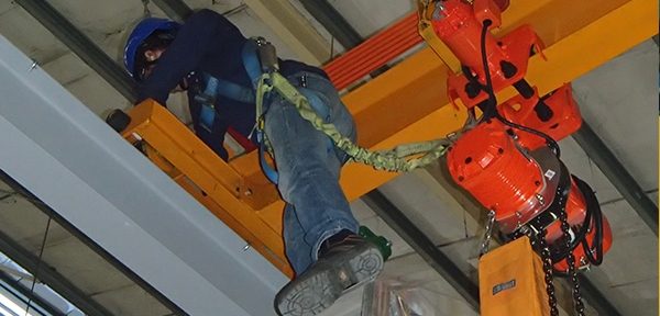 A technician checking a chain hoist and rigging assembly mounted on an overhead crane beam